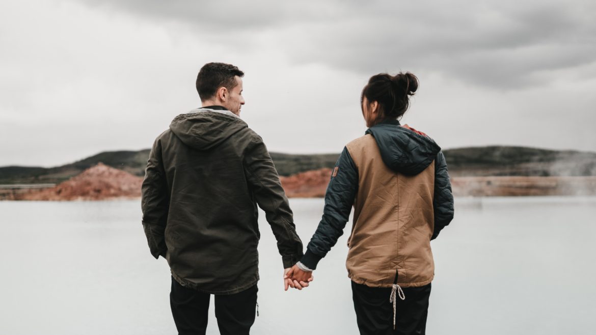 Two people looking at each other and holding hands in front of a lake.
